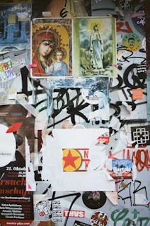 A collage of various posters, stickers, and graffiti layered over one another. Religious icons depicting the Virgin Mary and child, and another of a standing Mary figure are prominently placed at the top. Below them are multiple layers of torn posters and vibrant stickers with various designs and colors, including a circular red and yellow star image. Numerous black graffiti scribbles are visible, creating a chaotic and urban aesthetic.