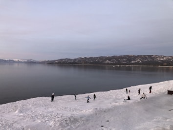 A snowy landscape with a large, calm body of water in the background and mountain ranges under a gray sky. Several people are engaged in activities on the snow-covered ground.