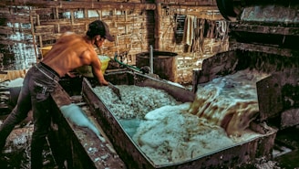 A shirtless man is working in an industrial setting, possibly involved in a manufacturing or processing task. He is handling materials in a large metal vat where a frothy liquid is being processed or drained. The environment is rustic, with wooden walls and a dimly lit atmosphere.