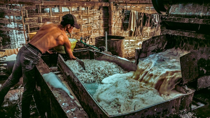 A shirtless man is working in an industrial setting, possibly involved in a manufacturing or processing task. He is handling materials in a large metal vat where a frothy liquid is being processed or drained. The environment is rustic, with wooden walls and a dimly lit atmosphere.