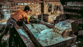 A shirtless man is working in an industrial setting, possibly involved in a manufacturing or processing task. He is handling materials in a large metal vat where a frothy liquid is being processed or drained. The environment is rustic, with wooden walls and a dimly lit atmosphere.