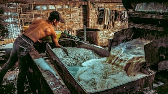 A shirtless man is working in an industrial setting, possibly involved in a manufacturing or processing task. He is handling materials in a large metal vat where a frothy liquid is being processed or drained. The environment is rustic, with wooden walls and a dimly lit atmosphere.