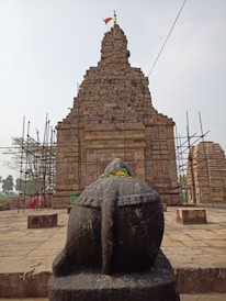 A stone statue of a Nandi bull is prominently displayed in the foreground, adorned with a garland of orange and yellow flowers. Behind the statue, a large ancient temple made of stacked stone bricks rises with scaffolding on either side. The temple has intricate carvings and a flag flying at the top, indicating restoration work might be ongoing. Several tall palm trees and a few people are in the background.