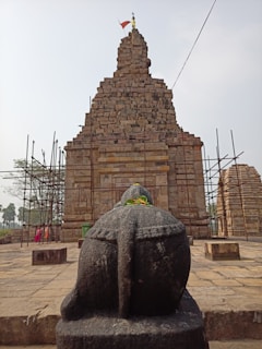 A stone statue of a Nandi bull is prominently displayed in the foreground, adorned with a garland of orange and yellow flowers. Behind the statue, a large ancient temple made of stacked stone bricks rises with scaffolding on either side. The temple has intricate carvings and a flag flying at the top, indicating restoration work might be ongoing. Several tall palm trees and a few people are in the background.