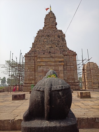 A stone statue of a Nandi bull is prominently displayed in the foreground, adorned with a garland of orange and yellow flowers. Behind the statue, a large ancient temple made of stacked stone bricks rises with scaffolding on either side. The temple has intricate carvings and a flag flying at the top, indicating restoration work might be ongoing. Several tall palm trees and a few people are in the background.
