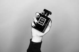 A hand with black nail polish is holding a black perfume bottle labeled 'Coco Noir Chanel Paris.' The image is in black and white, showcasing a minimalist and elegant aesthetic.