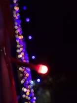 A close-up of a glowing red light in the foreground with a soft focus. In the background, colorful, out-of-focus lights in shades of blue, purple, and yellow create a bokeh effect against a dark setting.
