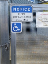 A metal gate with a sign instructing to keep it closed at all times. The sign is blue and white, featuring a notice and a wheelchair accessibility symbol. Chains are visible securing the gate, which has a grid-like metal pattern.