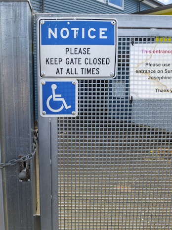 A metal gate with a sign instructing to keep it closed at all times. The sign is blue and white, featuring a notice and a wheelchair accessibility symbol. Chains are visible securing the gate, which has a grid-like metal pattern.