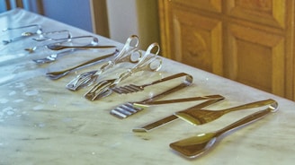 A collection of various silver kitchen utensils is neatly arranged on a white marble countertop. The assortment includes tongs, spatulas, and forks, each displayed in an orderly fashion. The background shows wooden cabinetry, adding warmth to the image.