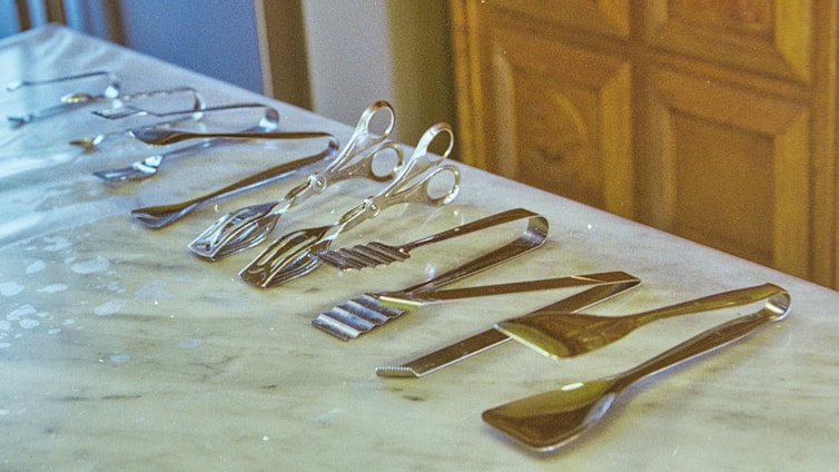 A collection of various silver kitchen utensils is neatly arranged on a white marble countertop. The assortment includes tongs, spatulas, and forks, each displayed in an orderly fashion. The background shows wooden cabinetry, adding warmth to the image.