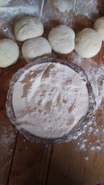 A wooden surface dusted with flour shows several pieces of dough arranged next to a silver platter filled with flour. The rustic setting and natural textures suggest a baking or bread-making activity.