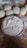 A wooden surface dusted with flour shows several pieces of dough arranged next to a silver platter filled with flour. The rustic setting and natural textures suggest a baking or bread-making activity.