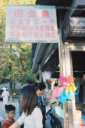 A small outdoor shop with a sign in Chinese offering goldfish. People, including children, are gathered around the counter where colorful toys like a multicolored pinwheel are displayed. The surrounding environment includes trees, suggesting a park setting.