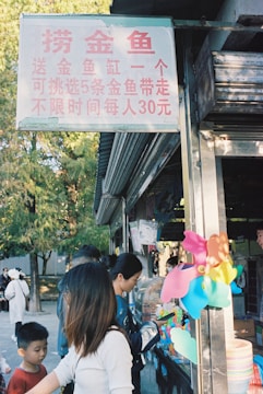 A small outdoor shop with a sign in Chinese offering goldfish. People, including children, are gathered around the counter where colorful toys like a multicolored pinwheel are displayed. The surrounding environment includes trees, suggesting a park setting.