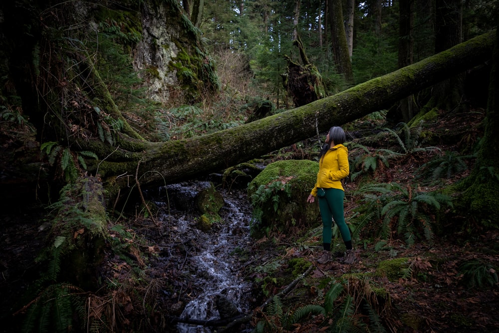 a woman in a yellow jacket standing in a forest next to a stream