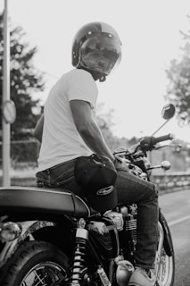 A person wearing a helmet and a white t-shirt is sitting on a motorcycle. The image is in black and white, creating a classic and timeless feel. The motorcycle's intricate mechanical details and the person's casual attire suggest a relaxed, biker lifestyle. Trees and a fence are visible in the background, indicating an outdoor setting.