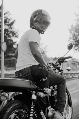 A person wearing a helmet and a white t-shirt is sitting on a motorcycle. The image is in black and white, creating a classic and timeless feel. The motorcycle's intricate mechanical details and the person's casual attire suggest a relaxed, biker lifestyle. Trees and a fence are visible in the background, indicating an outdoor setting.
