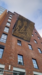 A red brick building with several windows and a large mural featuring stylized figures engaged in an activity involving teamwork or labor. The mural is set against a sky that is partially cloudy.