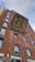 A red brick building with several windows and a large mural featuring stylized figures engaged in an activity involving teamwork or labor. The mural is set against a sky that is partially cloudy.