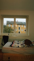 A bedroom with a window overlooking an orange apartment building. The room contains an unmade bed with a colorful, striped comforter and a brown backpack placed on it. A chair is positioned near the bed. Items are scattered on the window sill and a partially visible radiator is underneath the window.