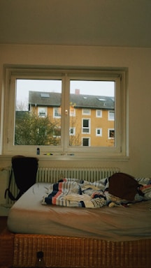 A bedroom with a window overlooking an orange apartment building. The room contains an unmade bed with a colorful, striped comforter and a brown backpack placed on it. A chair is positioned near the bed. Items are scattered on the window sill and a partially visible radiator is underneath the window.