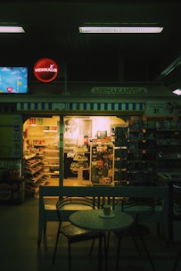 A dimly lit café inside a convenience store features a small round table with a cup on it in the foreground. Shelves filled with snacks and beverages are visible through the store's glass doors. A neon sign and various advertisements are displayed above and around the entrance.