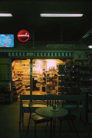 A dimly lit café inside a convenience store features a small round table with a cup on it in the foreground. Shelves filled with snacks and beverages are visible through the store's glass doors. A neon sign and various advertisements are displayed above and around the entrance.