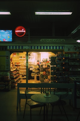 A dimly lit caf&eacute; inside a convenience store features a small round table with a cup on it in the foreground. Shelves filled with snacks and beverages are visible through the store's glass doors. A neon sign and various advertisements are displayed above and around the entrance.