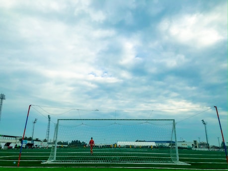 A wide view of a soccer field under a cloudy sky, featuring a soccer goal in the foreground. A goalkeeper in a red uniform stands by the goal, while other players are visible in the background, suggesting a soccer match is in progress. Floodlights surround the field, and some buildings can be seen in the distance.