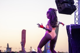 A performer with long curly hair wearing a colorful costume and red gloves is dancing on stage. The event seems to take place outdoors during sunset with sound equipment visible and flags or banners in the background, reflecting a lively and energetic atmosphere.