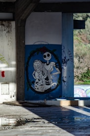 A piece of graffiti art featuring two cartoon characters, one with a skeleton-like appearance and the other with long hair, placed inside an oval frame with floral decorations at the top. The artwork is on a wall in an abandoned or dilapidated building with visible graffiti and exposed concrete. Debris and water puddles can be seen on the tiled floor.