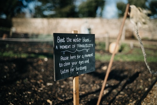 A wooden sign in a garden setting with text encouraging people to pause and enjoy tranquility. The sign is surrounded by natural elements like soil and plants, with blurred greenery in the background.