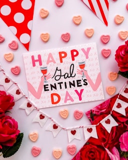 A festive and colorful arrangement featuring a card that reads 'Happy Galentine's Day,' surrounded by heart-shaped candy with messages and vibrant pink and red roses. There's a decorative garland with white and red heart flags, and the whole scene is set against a light pink background with polka dotted and striped patterns.