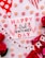A festive and colorful arrangement featuring a card that reads 'Happy Galentine's Day,' surrounded by heart-shaped candy with messages and vibrant pink and red roses. There's a decorative garland with white and red heart flags, and the whole scene is set against a light pink background with polka dotted and striped patterns.