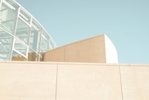 A modern architectural structure composed of glass and concrete elements. The building features a prominent glass section with a metal framework, reflecting a minimalist and contemporary design. The color palette is soft and muted, with light beige and pale blue tones dominating the scene, suggesting a peaceful and airy atmosphere.
