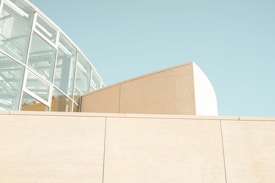 A modern architectural structure composed of glass and concrete elements. The building features a prominent glass section with a metal framework, reflecting a minimalist and contemporary design. The color palette is soft and muted, with light beige and pale blue tones dominating the scene, suggesting a peaceful and airy atmosphere.