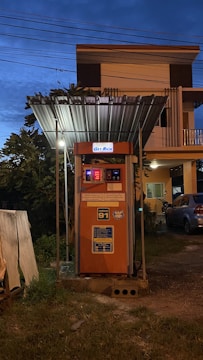 A gas pump station is situated in front of a house under a metal canopy. The structure is lit with several lights, creating a contrast with the evening sky. A vehicle and a motorcycle can be seen parked nearby.