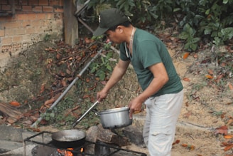 A man is outdoors cooking with a pot and pan over an open fire. He is wearing a green shirt and white pants, and appears focused on preparing food. The background includes a brick wall and lush greenery with fallen leaves around.