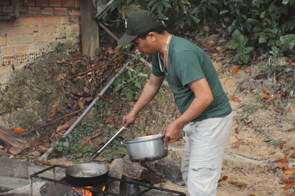 A man is outdoors cooking with a pot and pan over an open fire. He is wearing a green shirt and white pants, and appears focused on preparing food. The background includes a brick wall and lush greenery with fallen leaves around.