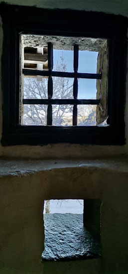 A view through two sets of windows, one on top of the other, each with a stone or concrete frame. The upper window features a grid-like design and offers a view of a partially snow-covered tree against a bright blue sky. The lower window is smaller, allowing a glimpse of a snowy landscape.