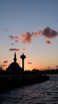 A silhouette of a mosque with a prominent dome and minaret stands against the backdrop of a sunset. The sky is adorned with scattered pink clouds, while birds are flying nearby. A large water tower is visible beside the mosque, and the foreground shows a body of water reflecting the twilight colors.