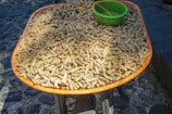 A large quantity of peanuts in their shells is displayed on a round orange tray. A green plastic bowl sits among the peanuts, contrasting with the rough cobblestone street in the background.