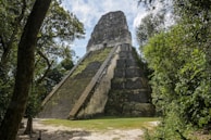 A large, ancient stone pyramid structure surrounded by lush green jungle vegetation. The pyramid is partly covered in moss and appears weathered by time. The sky above is partly cloudy.