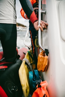 Two people wearing wetsuits are standing on a boat, holding onto a metal railing. Various colorful bags and safety equipment are attached to the boat's side.