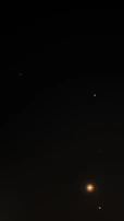 A dark night sky scattered with a few bright celestial bodies, including distant stars and what appears to be a planet or moon with a glowing aura. The prominence of the celestial object at the bottom right creates a sense of depth and distance.