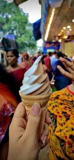 A relaxed crowd enjoying a day out, with a hand holding a soft-serve ice cream cone that swirls chocolate and vanilla flavors. The background is busy with people and the hint of a fair or outdoor event ambiance, featuring colorful clothes and blurred lights.