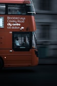 An orange double-decker bus in motion with destination details like Blackbird Leys, Cowley Road, city centre, and rail station displayed on its side. The focal point is the bus driver visible through the large window. The background appears blurred, emphasizing the bus's movement.