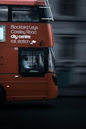 An orange double-decker bus in motion with destination details like Blackbird Leys, Cowley Road, city centre, and rail station displayed on its side. The focal point is the bus driver visible through the large window. The background appears blurred, emphasizing the bus's movement.