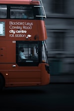 An orange double-decker bus in motion with destination details like Blackbird Leys, Cowley Road, city centre, and rail station displayed on its side. The focal point is the bus driver visible through the large window. The background appears blurred, emphasizing the bus's movement.